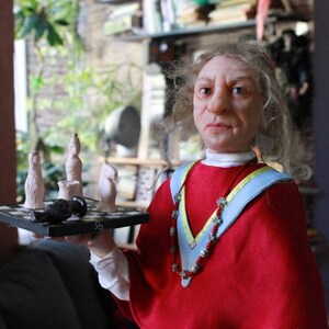 May include: A doll with long white hair wearing a red robe with a blue and yellow sash and a beaded necklace. The doll is holding a small chessboard with white chess pieces.