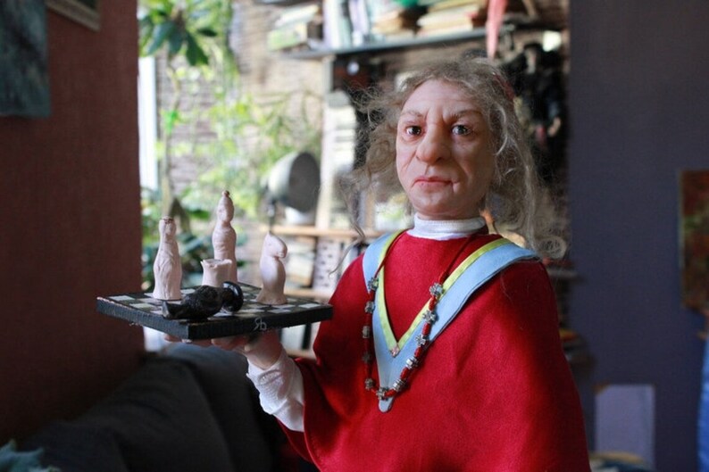 May include: A doll with long white hair wearing a red robe with a blue and yellow sash and a beaded necklace. The doll is holding a small chessboard with white chess pieces.
