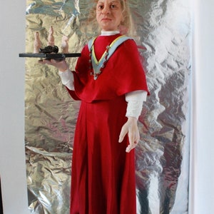 May include: A woman wearing a red robe and a blue and yellow sash holds a tray with a small black object and several white ceramic figures. She is standing on a stack of books.