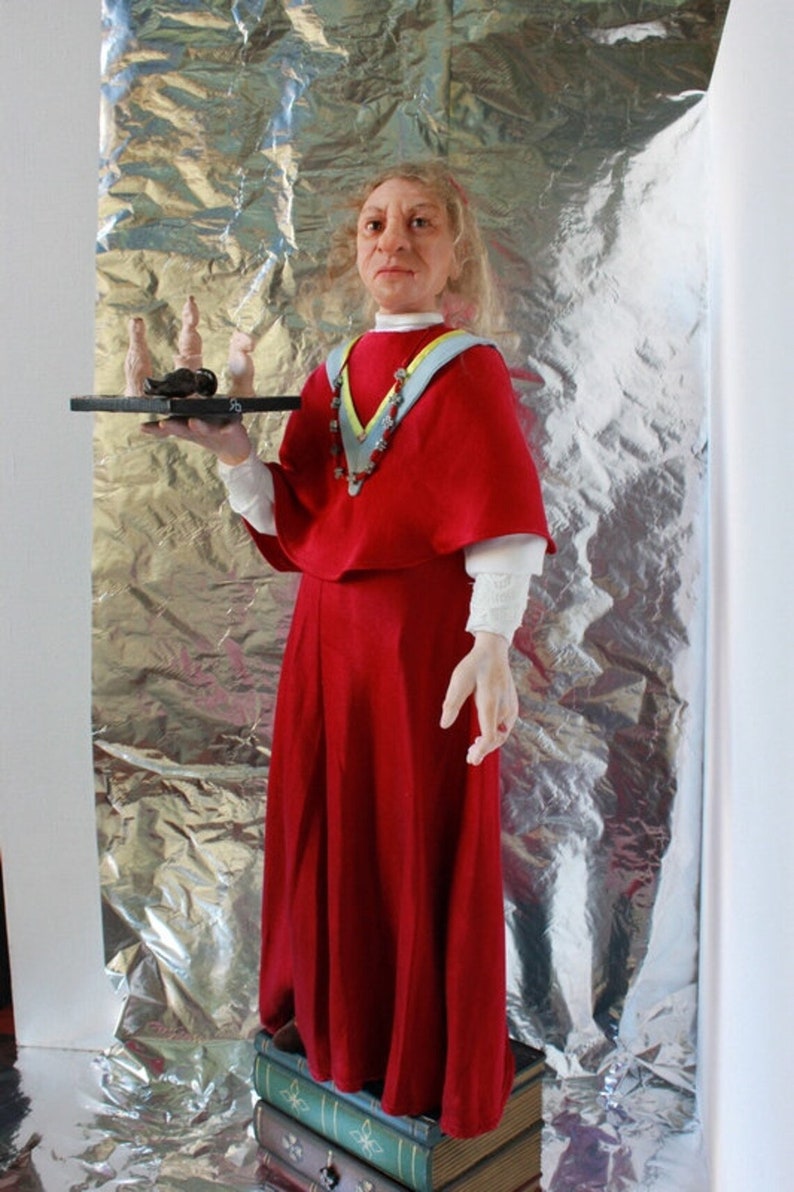 May include: A woman wearing a red robe and a blue and yellow sash holds a tray with a small black object and several white ceramic figures. She is standing on a stack of books.
