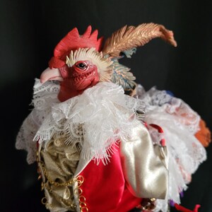 Pode incluir: Uma figura de galo vermelho com um colar de renda branca, uma corrente dourada e uma cauda de penas vermelhas e brancas. O galo est&aacute; vestindo uma jaqueta de cetim vermelha e branca.