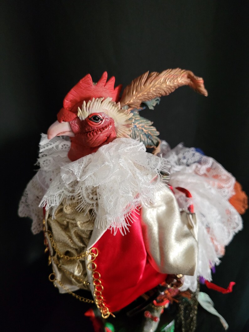 Pode incluir: Uma figura de galo vermelho com um colar de renda branca, uma corrente dourada e uma cauda de penas vermelhas e brancas. O galo est&aacute; vestindo uma jaqueta de cetim vermelha e branca.