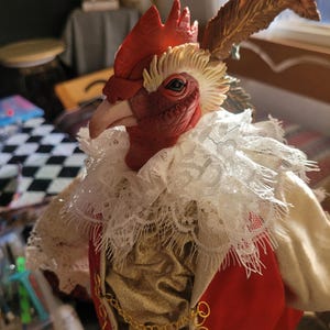 Pode incluir: Uma figura de galo vermelho usando um colar de renda branca, um colete dourado e uma jaqueta vermelha. O galo tem uma corrente dourada em volta do pesco&ccedil;o e uma pena marrom na cabe&ccedil;a.