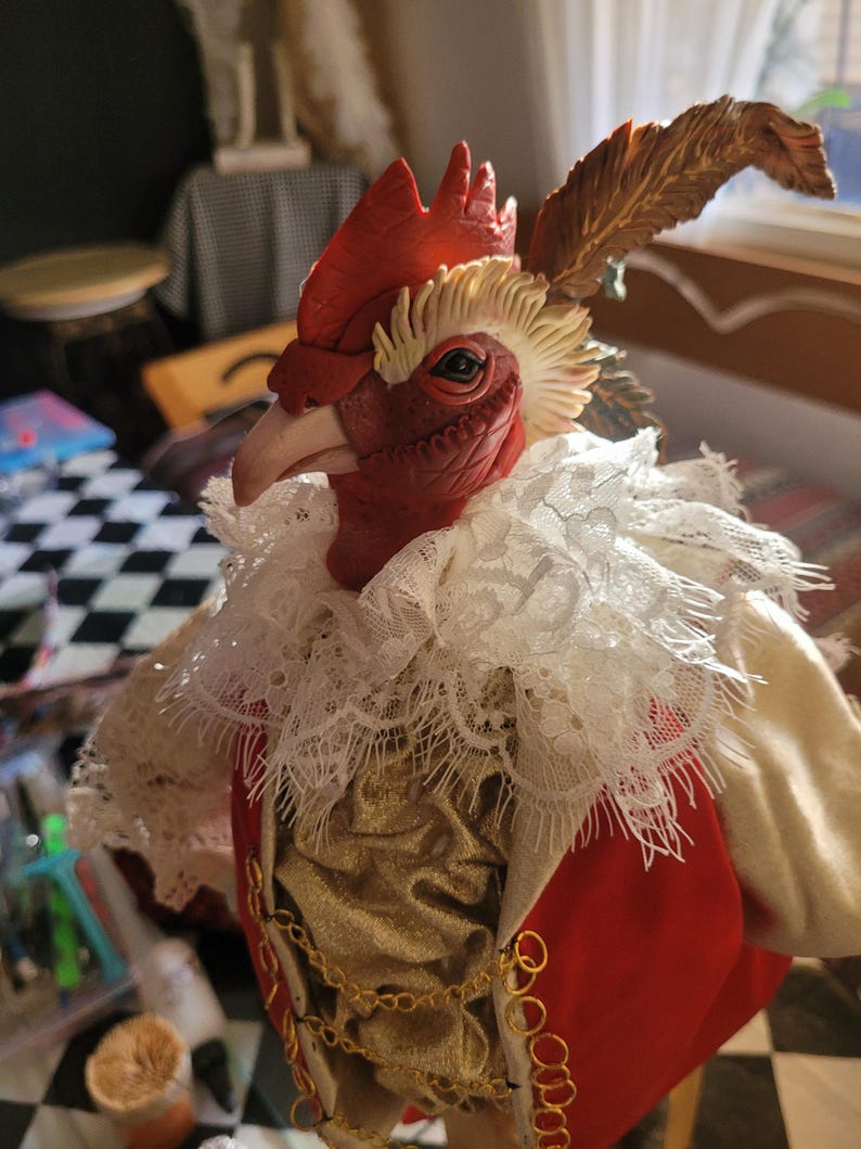 Pode incluir: Uma figura de galo vermelho usando um colar de renda branca, um colete dourado e uma jaqueta vermelha. O galo tem uma corrente dourada em volta do pesco&ccedil;o e uma pena marrom na cabe&ccedil;a.