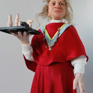 May include: A doll wearing a red dress and a blue and yellow sash with a beaded necklace. The doll is holding a tray with small white figurines.