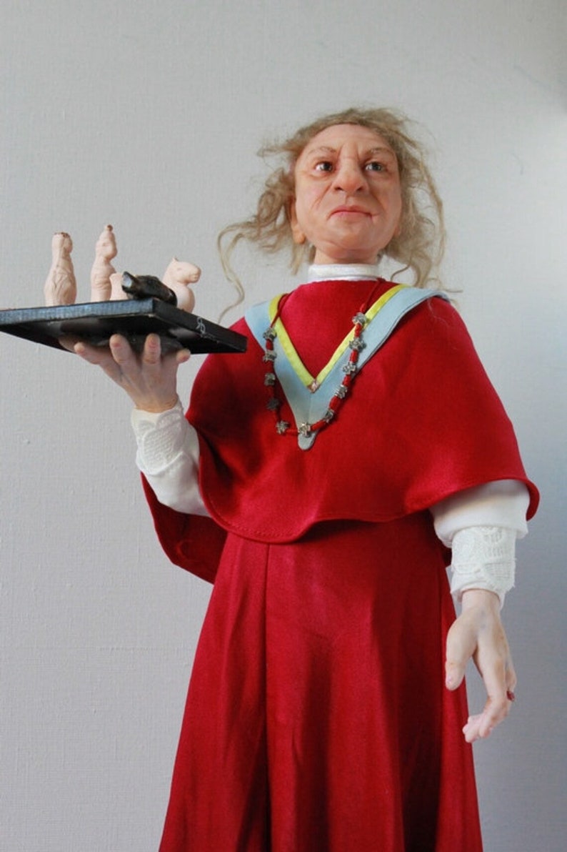 May include: A doll wearing a red dress and a blue and yellow sash with a beaded necklace. The doll is holding a tray with small white figurines.