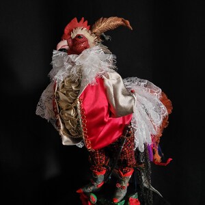 Pode incluir: Uma figura de galo vermelho vestindo um colar de renda branca, uma jaqueta vermelha com detalhes dourados e cal&ccedil;as com padr&atilde;o preto e vermelho. O galo est&aacute; em p&eacute; sobre uma base verde e vermelha com flores.