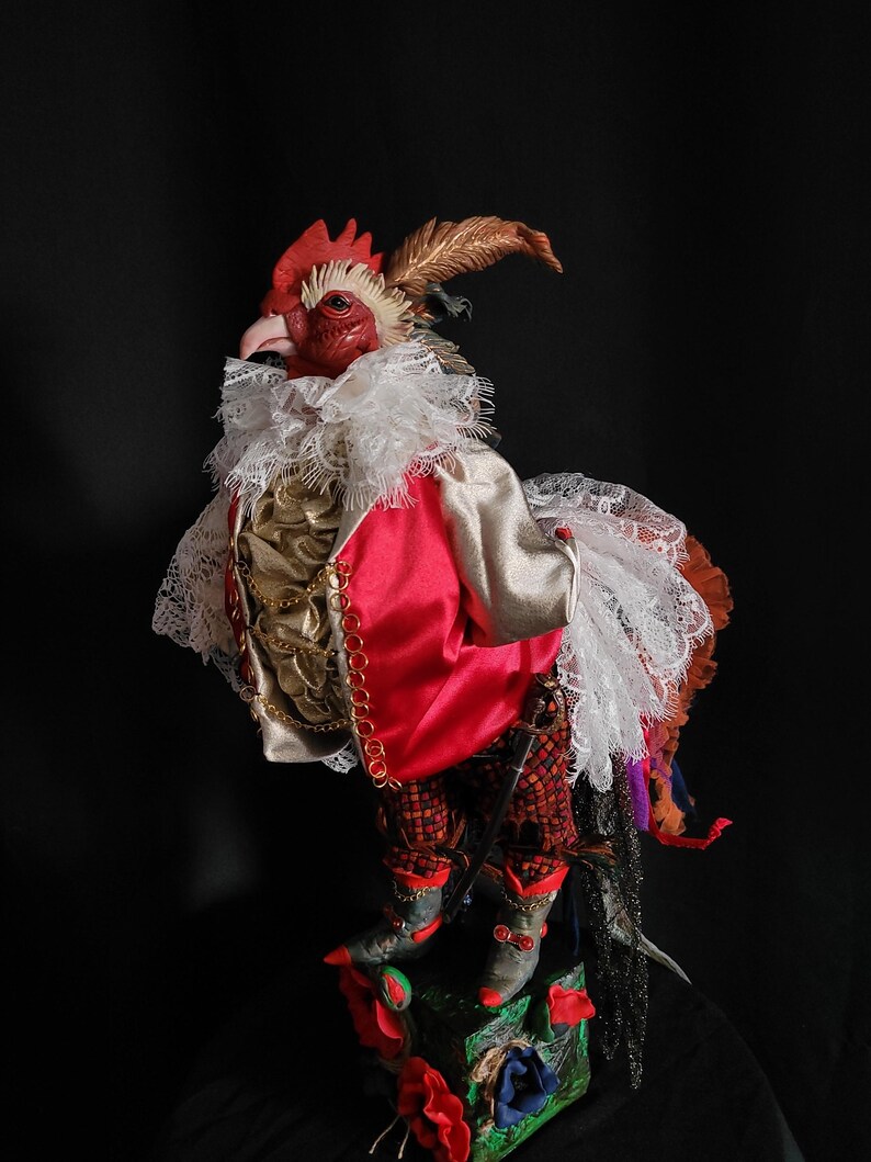 Pode incluir: Uma figura de galo vermelho vestindo um colar de renda branca, uma jaqueta vermelha com detalhes dourados e cal&ccedil;as com padr&atilde;o preto e vermelho. O galo est&aacute; em p&eacute; sobre uma base verde e vermelha com flores.