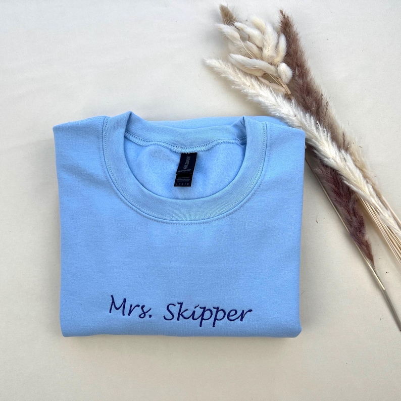 K&ouml;nnte beinhalten: Hellblaues Sweatshirt mit Rundhalsausschnitt und dem gestickten Text "Mrs. Skipper" in dunkelblauer Schrift. Das Sweatshirt ist ordentlich gefaltet und liegt neben getrockneten Zierpflanzen.