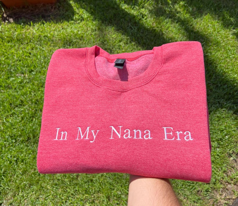 K&ouml;nnte beinhalten: Ein rotes Sweatshirt mit dem wei&szlig;en Schriftzug "In My Nana Era". Das Sweatshirt mit Rundhalsausschnitt ist aus weichem Material gefertigt und wird auf einem gr&uuml;nen, grasbewachsenen Hintergrund pr&auml;sentiert.