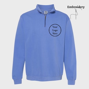 May include: A light blue quarter-zip sweatshirt with a collar and a zipper. The sweatshirt has a circular logo area that says "Your Logo Here". The word "Embroidery" is written in the upper right corner.