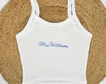Op de afbeelding: Witte tanktop met dunne bandjes. De tekst "Mrs. Williams" is in blauw script op de voorkant geborduurd. De top wordt getoond op een geweven, ronde mat.