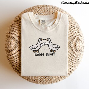May include: A cream colored t-shirt with an embroidered design of two geese facing each other with the text "Goose Bumps" underneath.