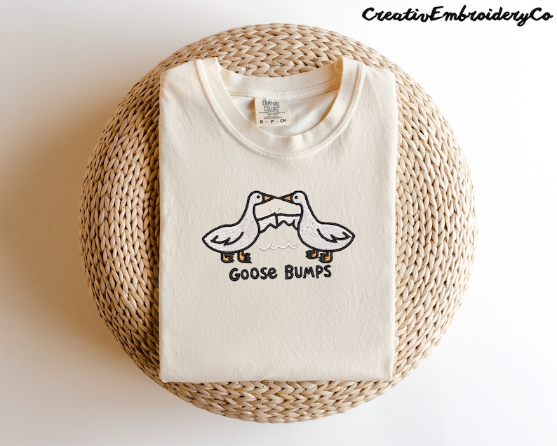 May include: A cream colored t-shirt with an embroidered design of two geese facing each other with the text "Goose Bumps" underneath.