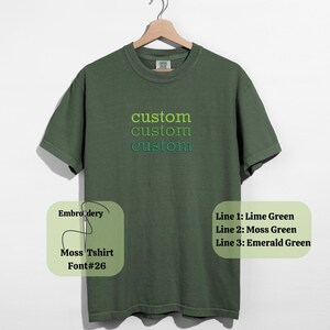 May include: A moss green t-shirt with the word "custom" embroidered in lime green, moss green, and emerald green. The shirt is displayed on a wooden hanger against a white background. The text "Moss Tshirt Font#26" is also visible.