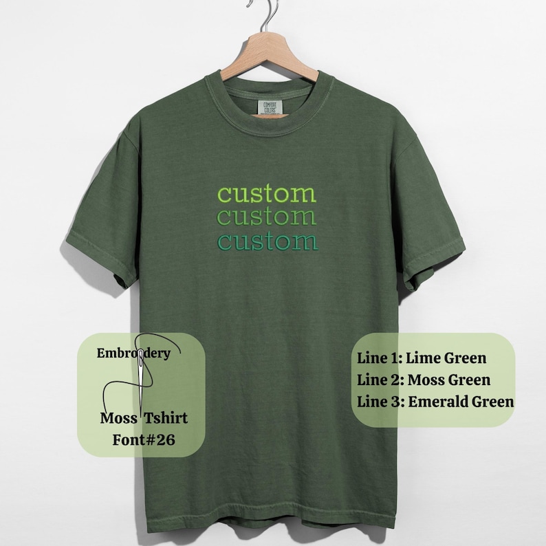 May include: A moss green t-shirt with the word "custom" embroidered in lime green, moss green, and emerald green. The shirt is displayed on a wooden hanger against a white background. The text "Moss Tshirt Font#26" is also visible.