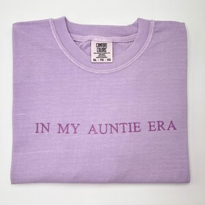 May include: A lavender Comfort Colors t-shirt, folded neatly, with the phrase "IN MY AUNTIE ERA" embroidered in a darker purple. The shirt has a crew neck and a tag visible at the neckline.