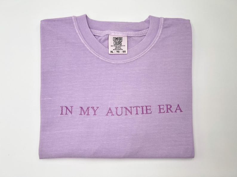 May include: A lavender Comfort Colors t-shirt, folded neatly, with the phrase "IN MY AUNTIE ERA" embroidered in a darker purple. The shirt has a crew neck and a tag visible at the neckline.
