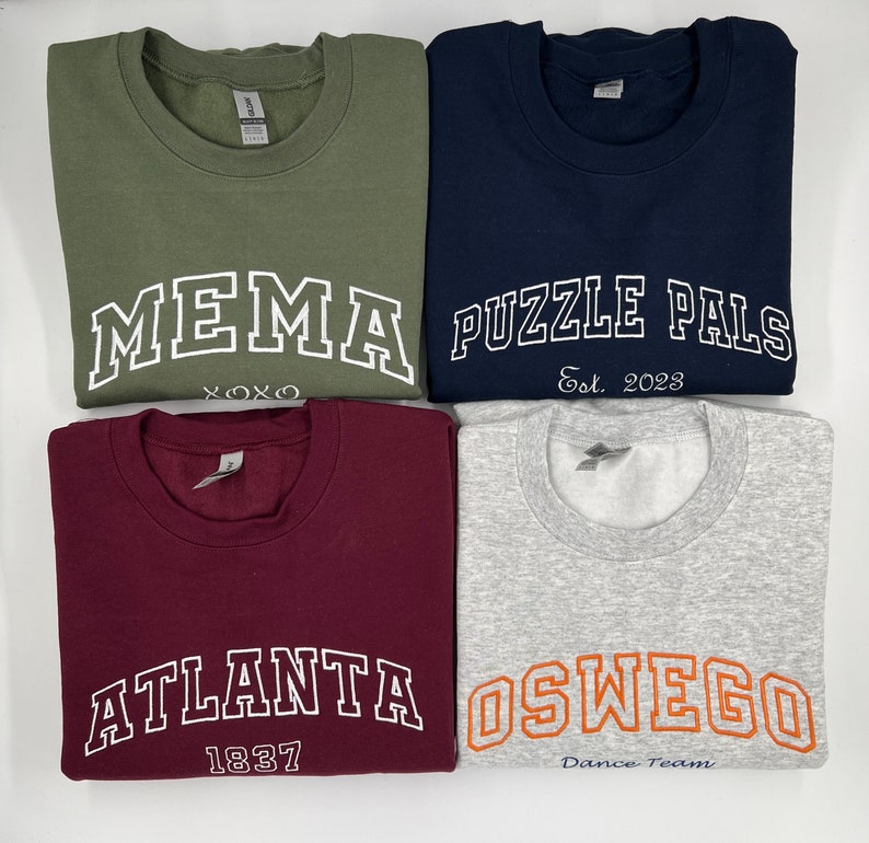 May include: Four sweatshirts with different designs and colors. The first sweatshirt is olive green with white text that says "MEMA XOXO". The second sweatshirt is navy blue with white text that says "PUZZLE PALS Est. 2023". The third sweatshirt is maroon with white text that says "ATLANTA 1837". The fourth sweatshirt is light gray with orange text that says "OSWEGO Dance Team".
