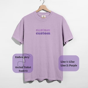 May include: A lilac-colored t-shirt with the word "custom" embroidered twice in lilac and purple. The shirt is hanging on a wooden hanger. The text "Embroidery, Orchid Tshirt, Font#3, Line 1: Lilac, Line 2: Purple" is also visible.