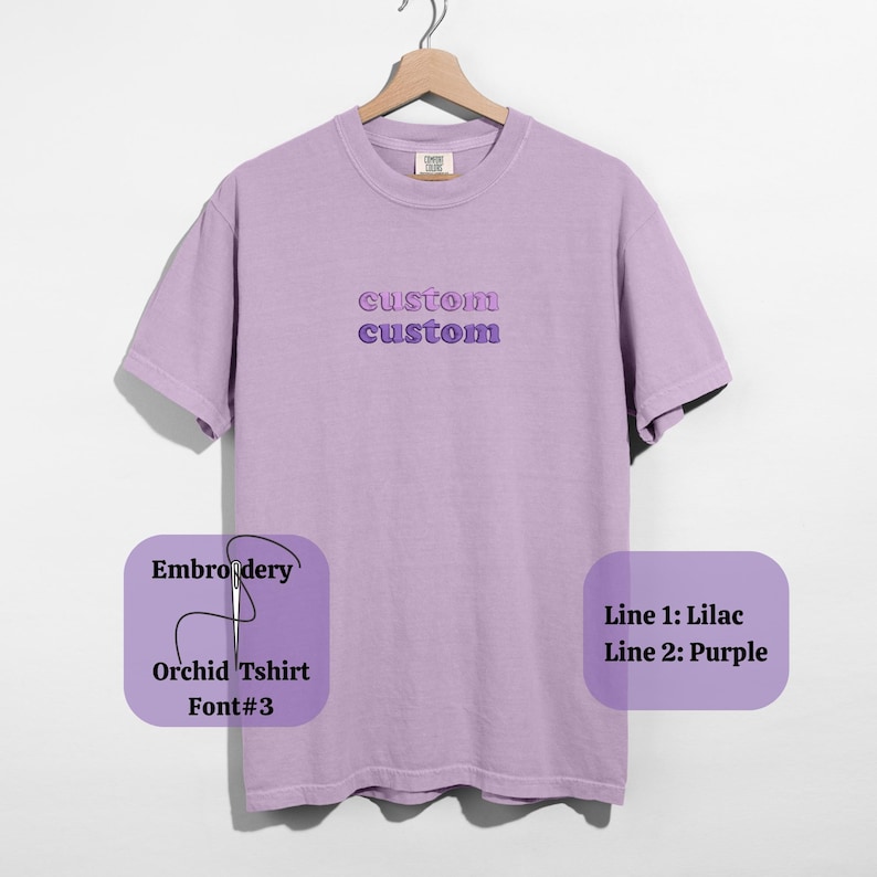 May include: A lilac-colored t-shirt with the word "custom" embroidered twice in lilac and purple. The shirt is hanging on a wooden hanger. The text "Embroidery, Orchid Tshirt, Font#3, Line 1: Lilac, Line 2: Purple" is also visible.