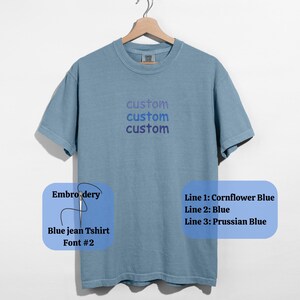 May include: A blue jean t-shirt with the word "custom" embroidered three times in blue thread. The embroidery colors are Cornflower Blue, Blue, and Prussian Blue. The shirt is hanging on a wooden hanger.