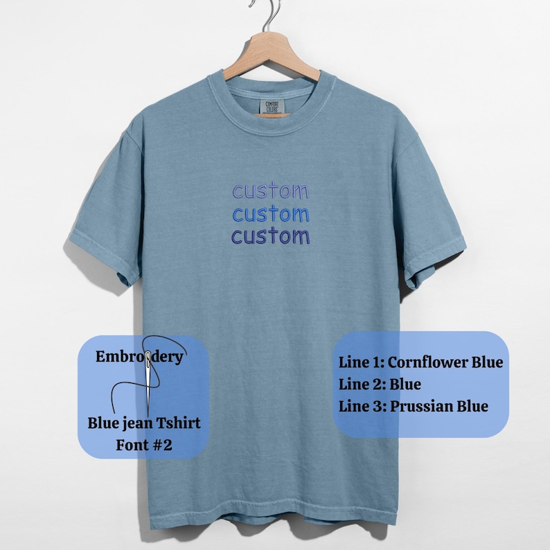 May include: A blue jean t-shirt with the word "custom" embroidered three times in blue thread. The embroidery colors are Cornflower Blue, Blue, and Prussian Blue. The shirt is hanging on a wooden hanger.