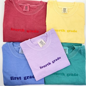 May include: A collection of Comfort Colors t-shirts in various colors, including red, yellow, blue, purple, and teal. Each shirt has "fourth grade" or "first grade" embroidered on the front. The shirts are folded and arranged on a white surface.