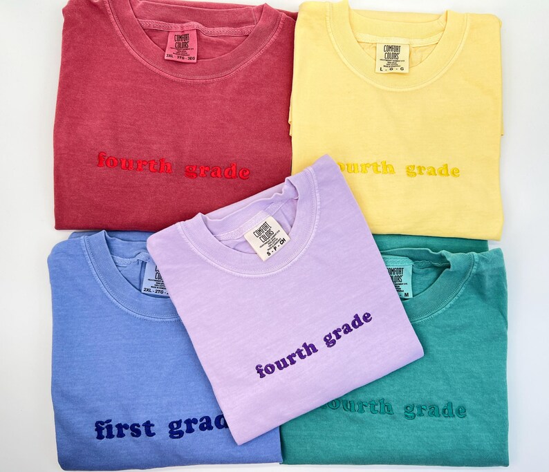 May include: A collection of Comfort Colors t-shirts in various colors, including red, yellow, blue, purple, and teal. Each shirt has "fourth grade" or "first grade" embroidered on the front. The shirts are folded and arranged on a white surface.