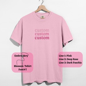 May include: A light pink t-shirt with the word "custom" embroidered three times in varying shades of pink. The shirt is hanging on a wooden hanger. The text "Embroidery, Blossom Tshirt, Font#7" is also visible.