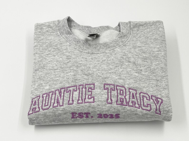 May include: Gray sweatshirt with purple embroidered text that reads "Auntie Tracy Est. 2023".