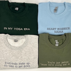 K&ouml;nnte beinhalten: Vier Sweatshirts mit Rundhalsausschnitt in verschiedenen Farben: Dunkelgr&uuml;n, Hellblau, Grau und Olivgr&uuml;n. Jedes Sweatshirt hat einen wei&szlig;en gestickten Text. Die Phrasen lauten: "IN MY YOGA ERA", "HEART WARRIOR MAMA", "Everybody wake up it's time to get down" und "You're just jealous 'cause we're young and in love".