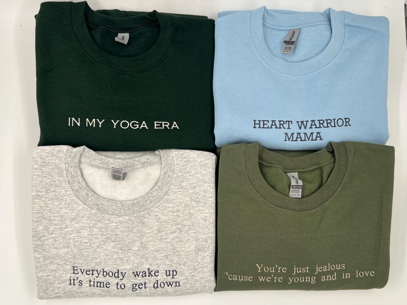 K&ouml;nnte beinhalten: Vier Sweatshirts mit Rundhalsausschnitt in verschiedenen Farben: Dunkelgr&uuml;n, Hellblau, Grau und Olivgr&uuml;n. Jedes Sweatshirt hat einen wei&szlig;en gestickten Text. Die Phrasen lauten: "IN MY YOGA ERA", "HEART WARRIOR MAMA", "Everybody wake up it's time to get down" und "You're just jealous 'cause we're young and in love".