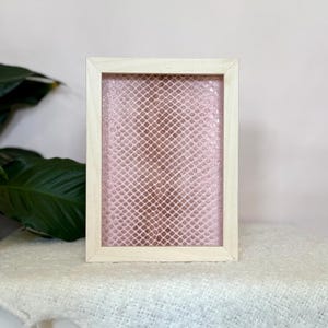 May include: A rectangular wooden frame with a pink, textured interior. The frame is made of light-coloured wood and sits on a white surface. The pink interior has a grid-like pattern.