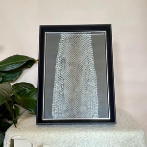 May include: A framed artwork with a snakeskin pattern in grey and white tones. The black frame has a white mount. The artwork is displayed on a white table, covered with a textured, off-white throw. The artwork is approximately 30 cm x 40 cm.