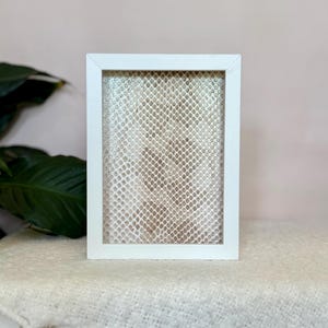 May include: A white rectangular picture frame with a snakeskin pattern inside. The frame is made of wood and is on a white surface. The background is a light pink colour.