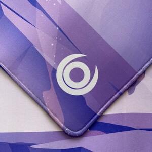 May include: Purple and white mousepad with a geometric design and a white circle logo in the center. The mousepad has a stitched edge.