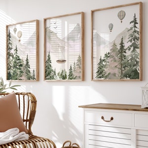May include: Three framed watercolour paintings of mountains and trees with hot air balloons in the sky. The paintings are hanging on a white wall above a white dresser with a brown top. A wicker chair with a brown cushion and a white throw blanket is in the foreground.