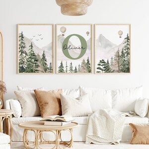 May include: Three watercolour prints featuring a mountain landscape with green trees, a light pink sky, and hot air balloons. The middle print features the letter "O" in green with the name "Oliver" written below it.