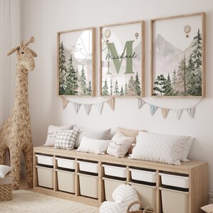May include: Three framed prints featuring a watercolour mountain and forest scene. The central print displays the letter "M" with the name "Michael" written beneath it. The prints are hung on a white wall above a wooden storage bench with white bins.