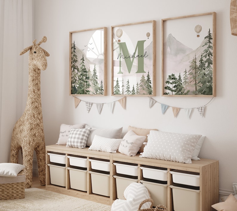 May include: Three framed prints featuring a watercolour mountain and forest scene. The central print displays the letter "M" with the name "Michael" written beneath it. The prints are hung on a white wall above a wooden storage bench with white bins.