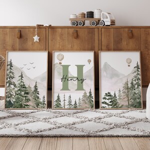 May include: Three framed watercolour prints featuring a mountain and forest scene. The centre print includes the letter H and the name Henry. The prints are displayed on a wooden shelf.