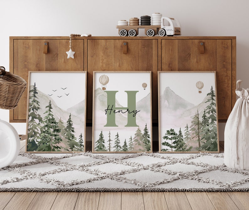 May include: Three framed watercolour prints featuring a mountain and forest scene. The centre print includes the letter H and the name Henry. The prints are displayed on a wooden shelf.