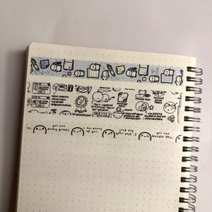 May include: A close-up of a notebook with a white background and black dots. The notebook has a spiral binding and is decorated with several washi tapes featuring cute cartoon characters and motivational phrases. The washi tapes are in various colours, including blue, pink, and yellow. The phrases on the washi tapes include "You are doing great!" and "I am proud of you."