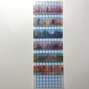 May include: A set of six washi tapes featuring a cityscape design. The tapes are arranged vertically on a gridded background. The tapes are translucent and feature a variety of colors, including blue, pink, and gold.