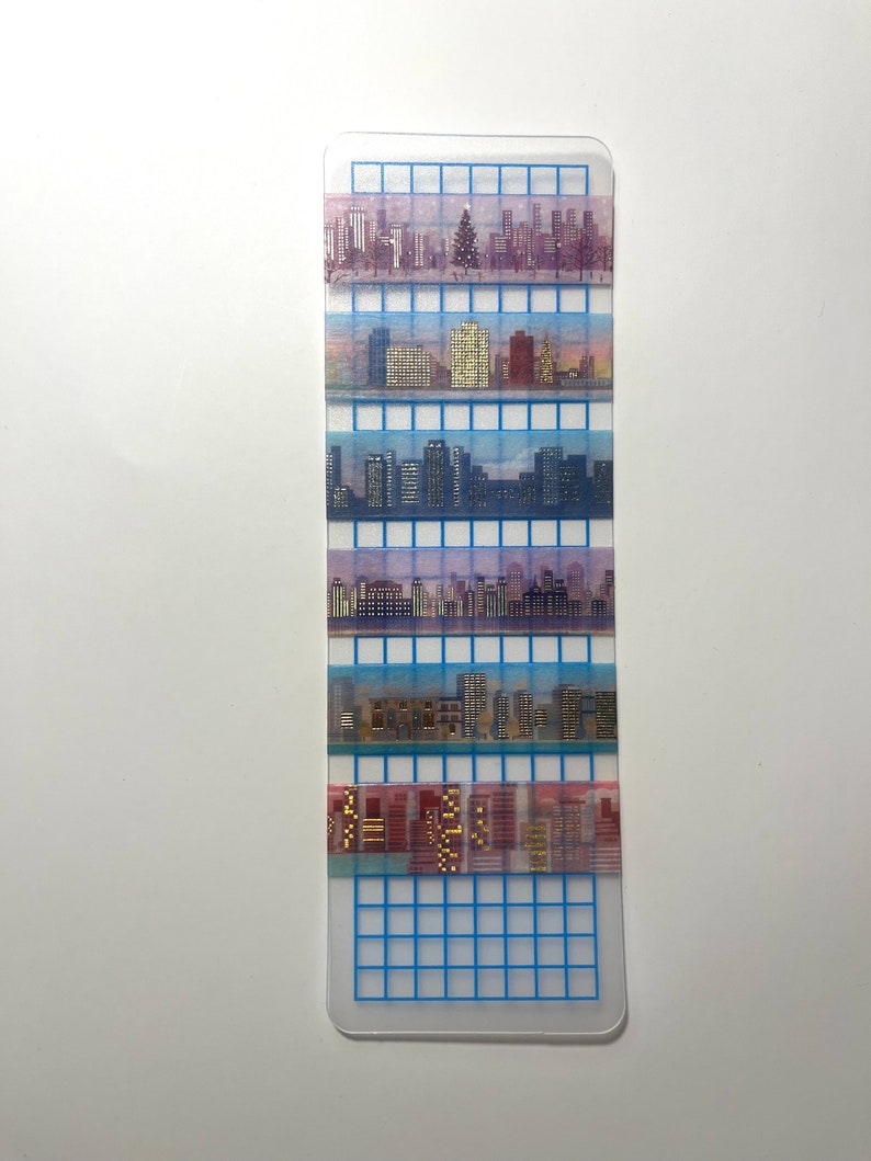 May include: A set of six washi tapes featuring a cityscape design. The tapes are arranged vertically on a gridded background. The tapes are translucent and feature a variety of colors, including blue, pink, and gold.