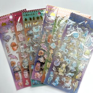May include: Four packs of clear plastic stickers with colorful illustrations of animals, plants, and objects. The stickers are arranged in a stack, with the top pack showing a blue and white cat with a pink nose and a pink bow. The stickers are labeled "NEKONI" in gold lettering.