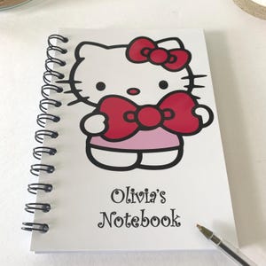 May include: White spiral-bound notebook with a Hello Kitty graphic, featuring a red bow and pink dress. The notebook is personalised with the text "Olivia's Notebook" in black script. A pen rests on the bottom right corner.
