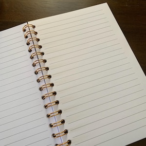 May include: Open notebook with lined white pages and a gold spiral binding. The notebook is open to a blank page, ready for writing or sketching. The lines are evenly spaced, and the paper appears smooth.