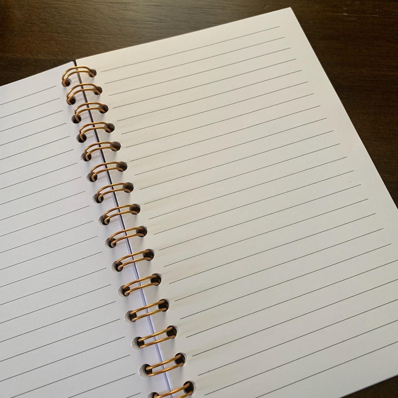 May include: Open notebook with lined white pages and a gold spiral binding. The notebook is open to a blank page, ready for writing or sketching. The lines are evenly spaced, and the paper appears smooth.
